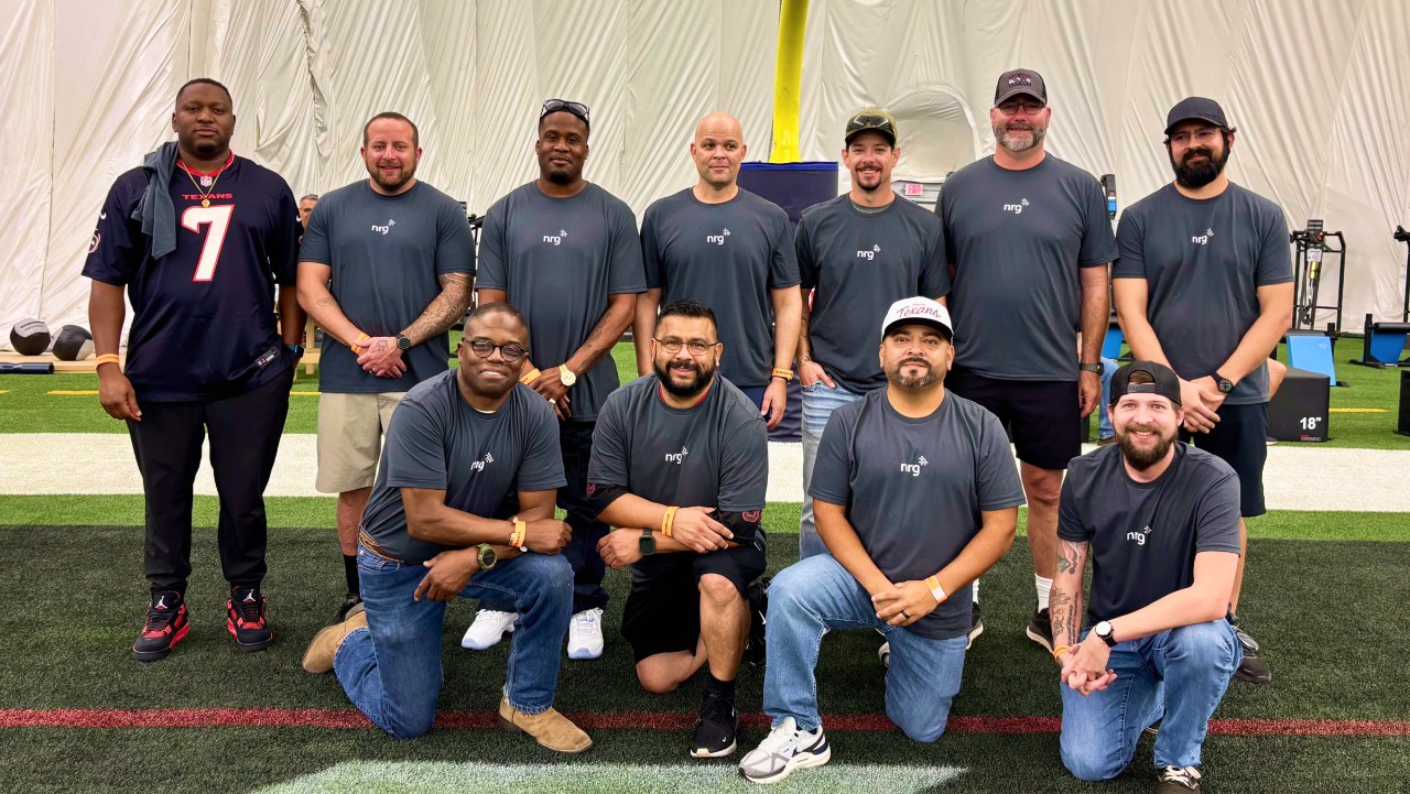 twelve men pose indoors on turf, most wearing matching gray shirts, some kneeling in front, others standing behind.