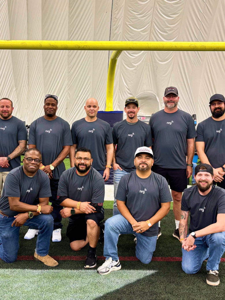 nine men in matching gray shirts pose and smile together under a yellow goalpost on indoor turf.