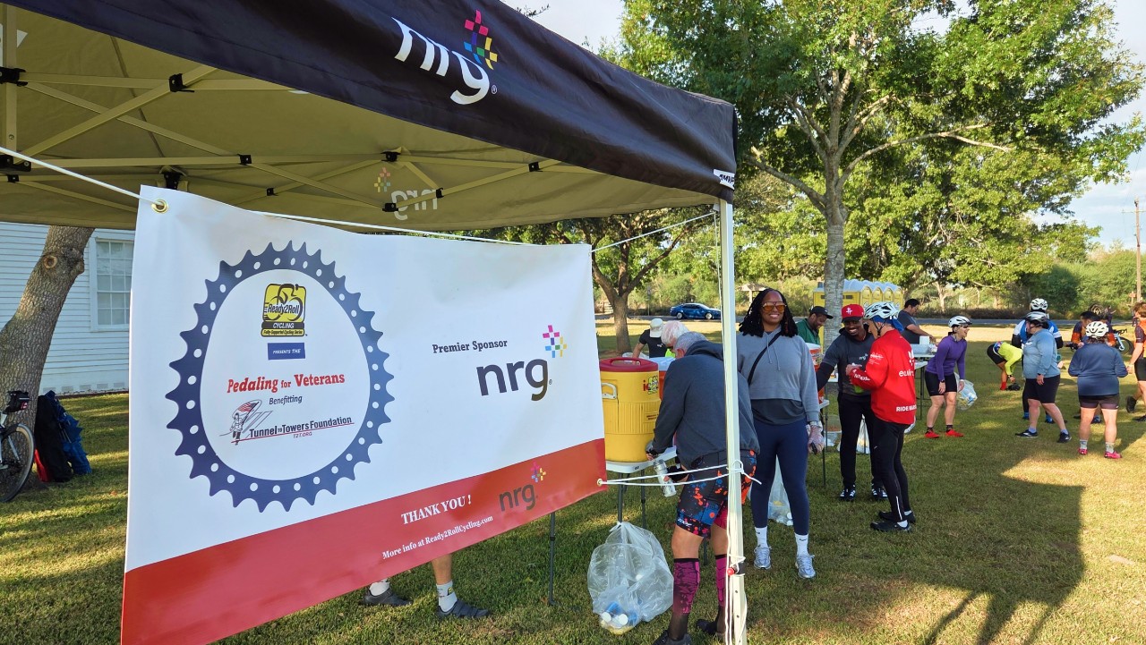 people gather near a tent with a Pedaling 4 Veterans banner at an outdoor event on a sunny day.