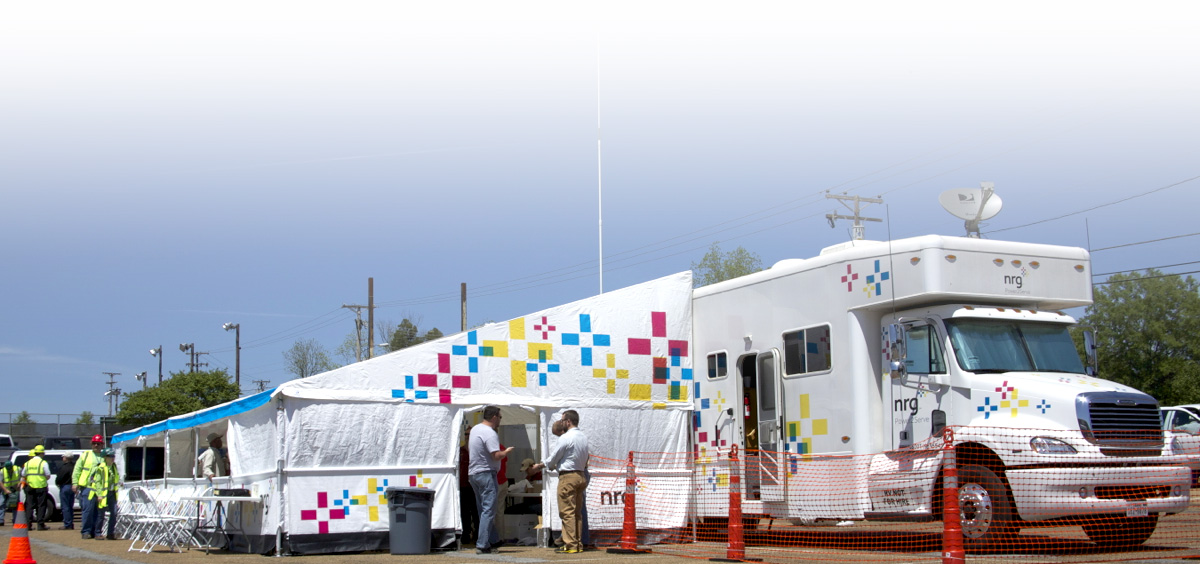 white tent and large truck with colorful square logo, surrounded by people and orange safety fencing.