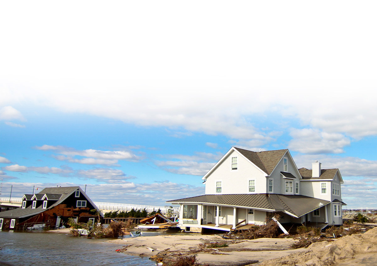 two damaged beach houses surrounded by sand and debris under a blue sky with clouds.