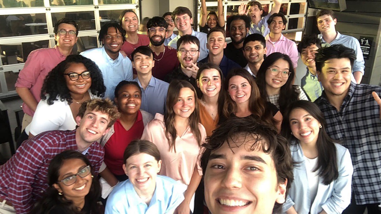 a large group of young adults smiling and posing for a selfie in an indoor setting with windows behind them.