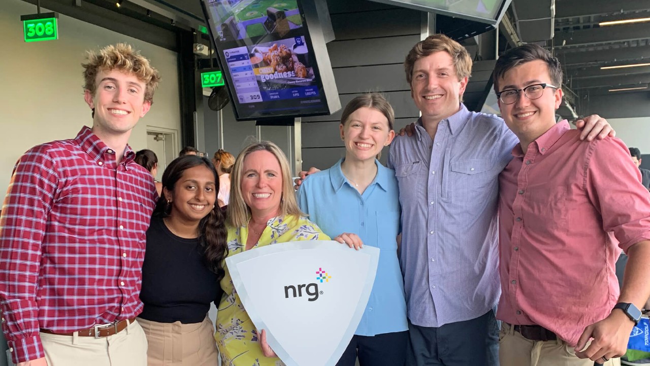 six people smiling and posing together indoors, one holding a white sign with NRG. written on it.