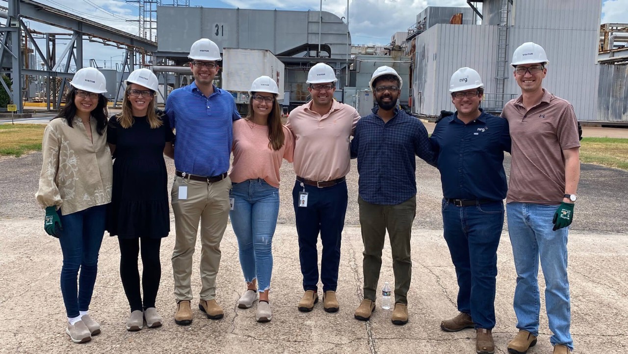 eight people in hard hats and casual clothes stand together and smile at an industrial site outdoors.