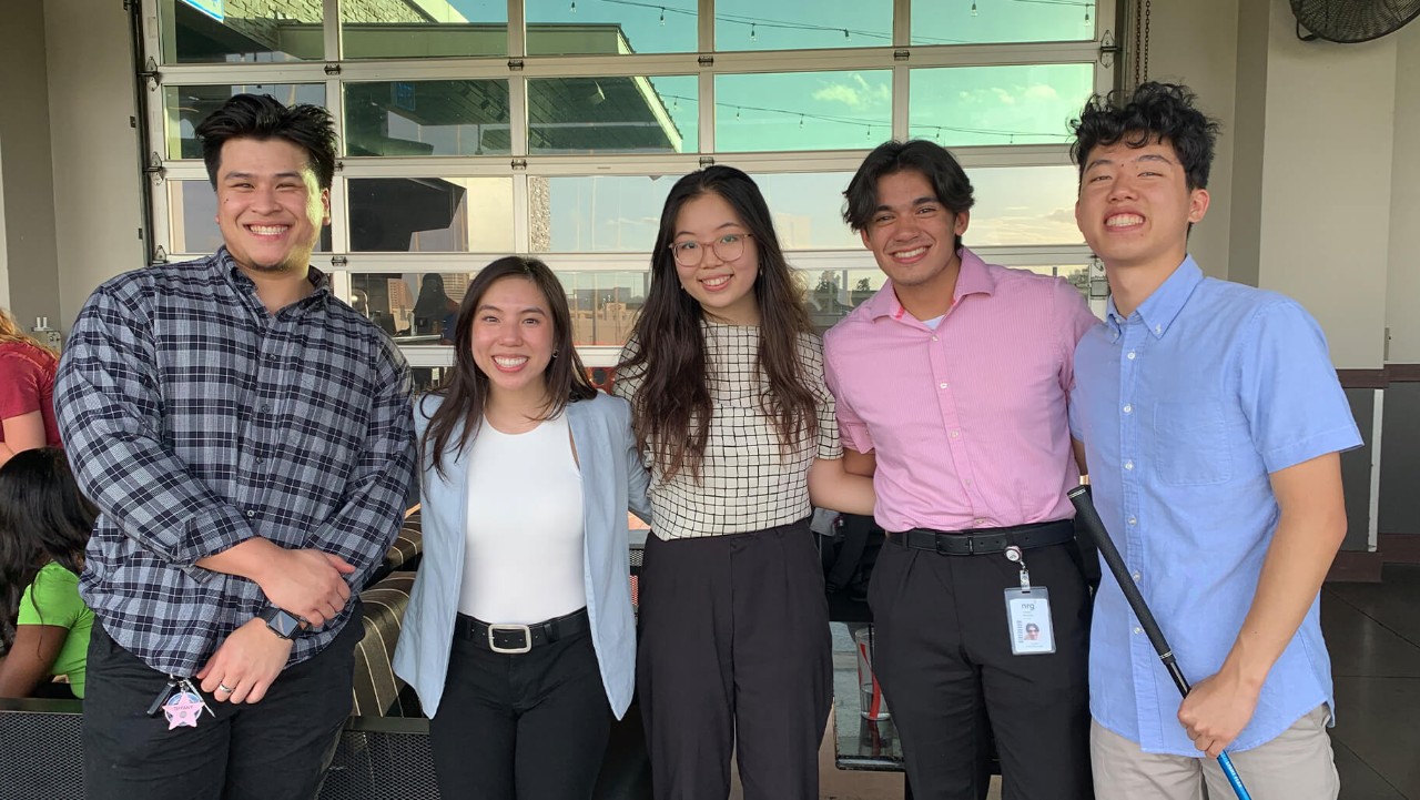 five young adults smiling and standing together indoors, dressed in business casual attire.