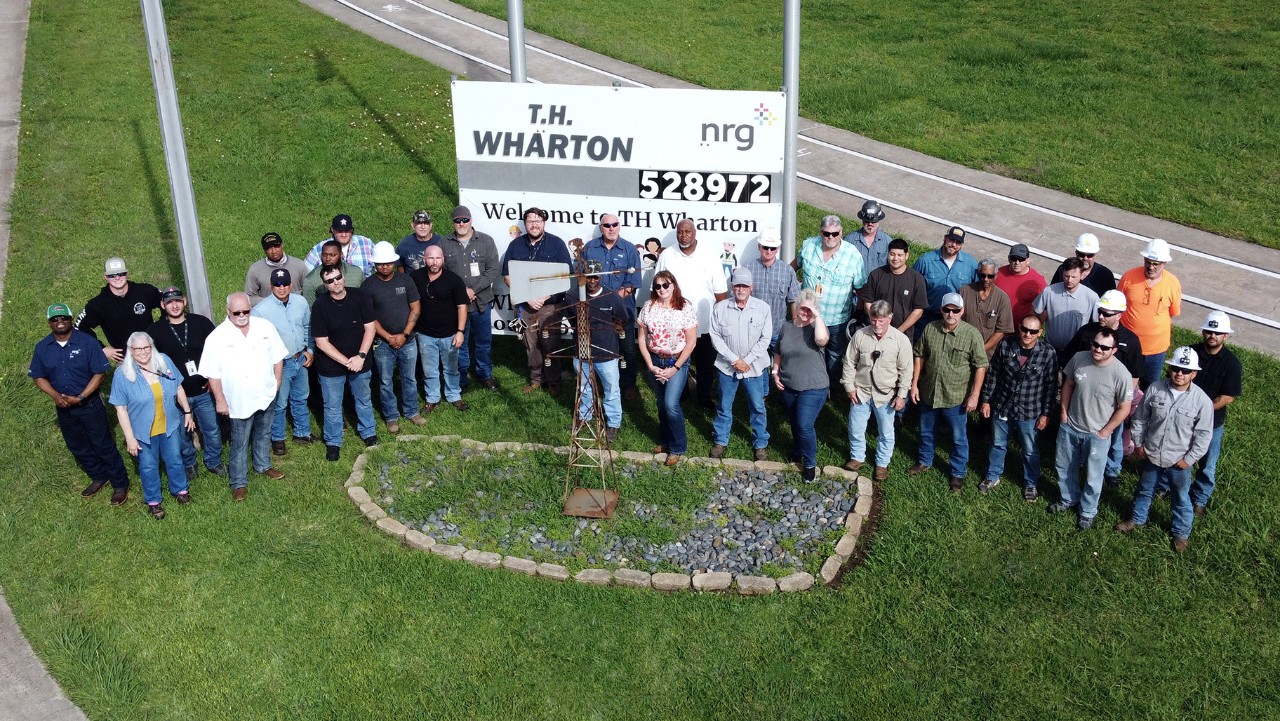 group of workers posing outdoors by a Welcome to T.H. Wharton NRG sign, aerial view.