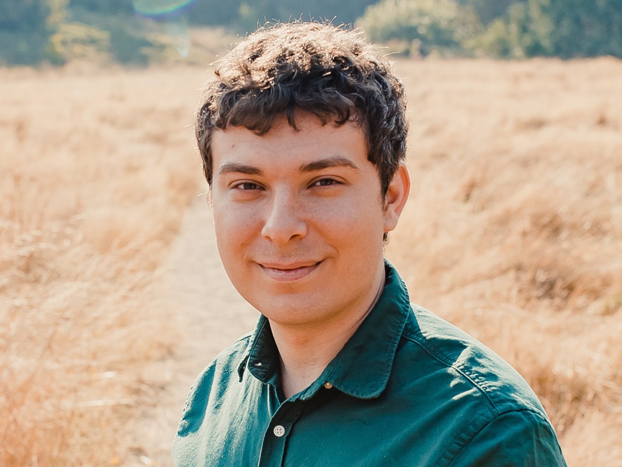 a person with short curly hair smiles outdoors in a dry, grassy field, wearing a green shirt.