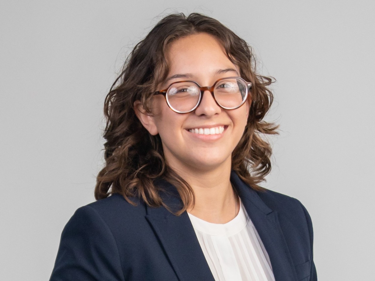 person with glasses and shoulder-length hair smiling, wearing a navy blazer and white shirt, against a gray background.