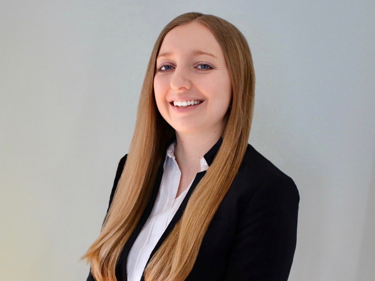 smiling woman with long straight hair, wearing a black blazer and white shirt, standing against a plain background.