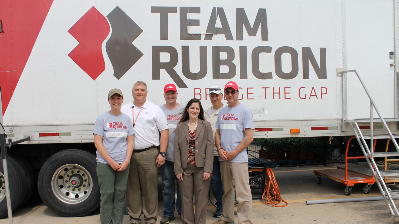 six people, some in Team Rubicon shirts, stand smiling in front of a Team Rubicon trailer.