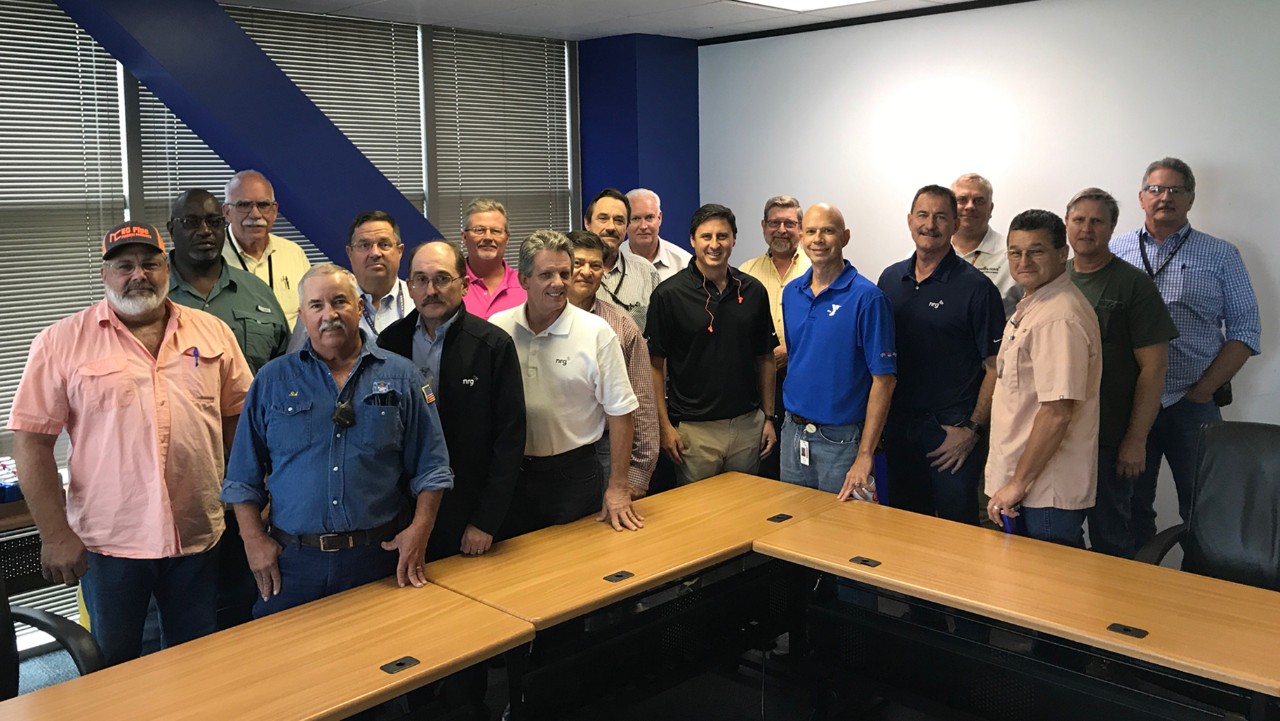 a group of men stand and smile together in a conference room behind wooden tables.