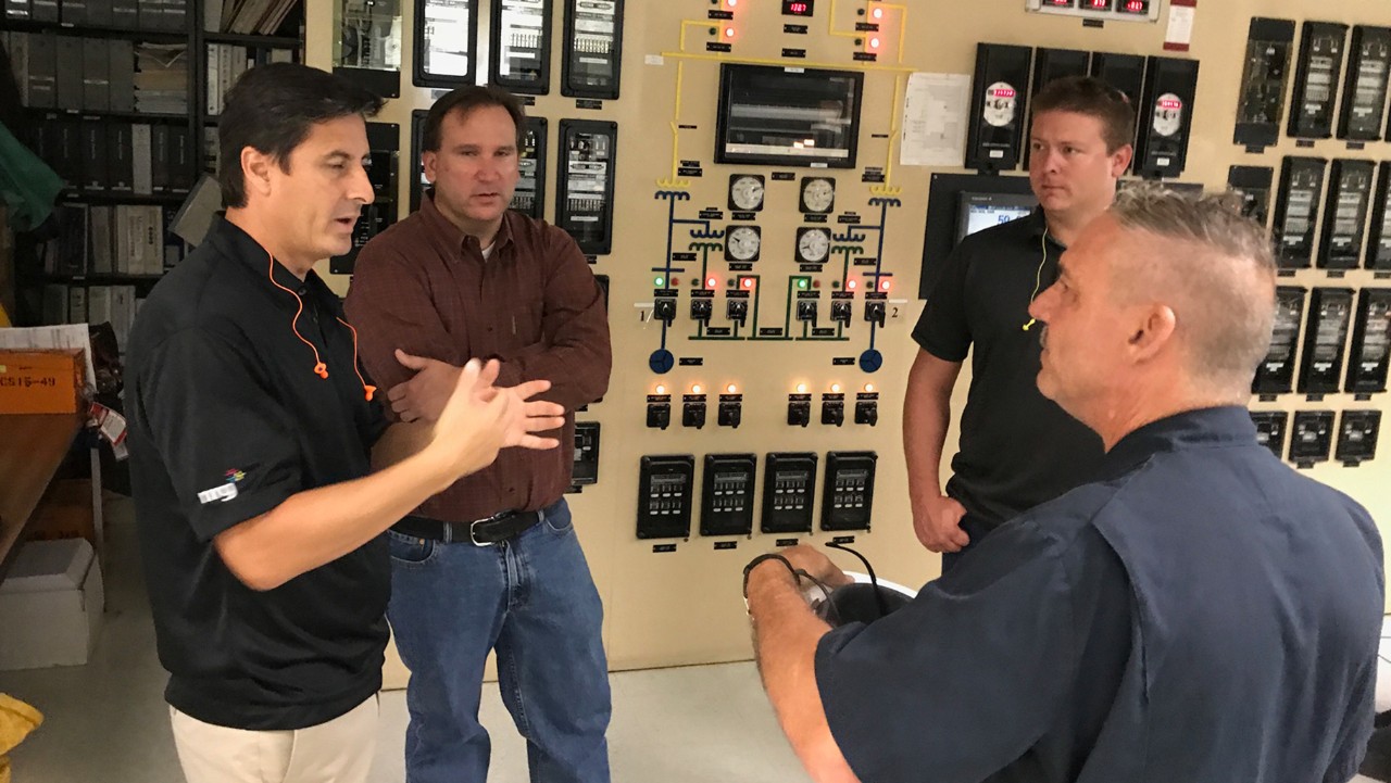 four men have a discussion in a control room filled with panels, switches, and monitoring equipment.