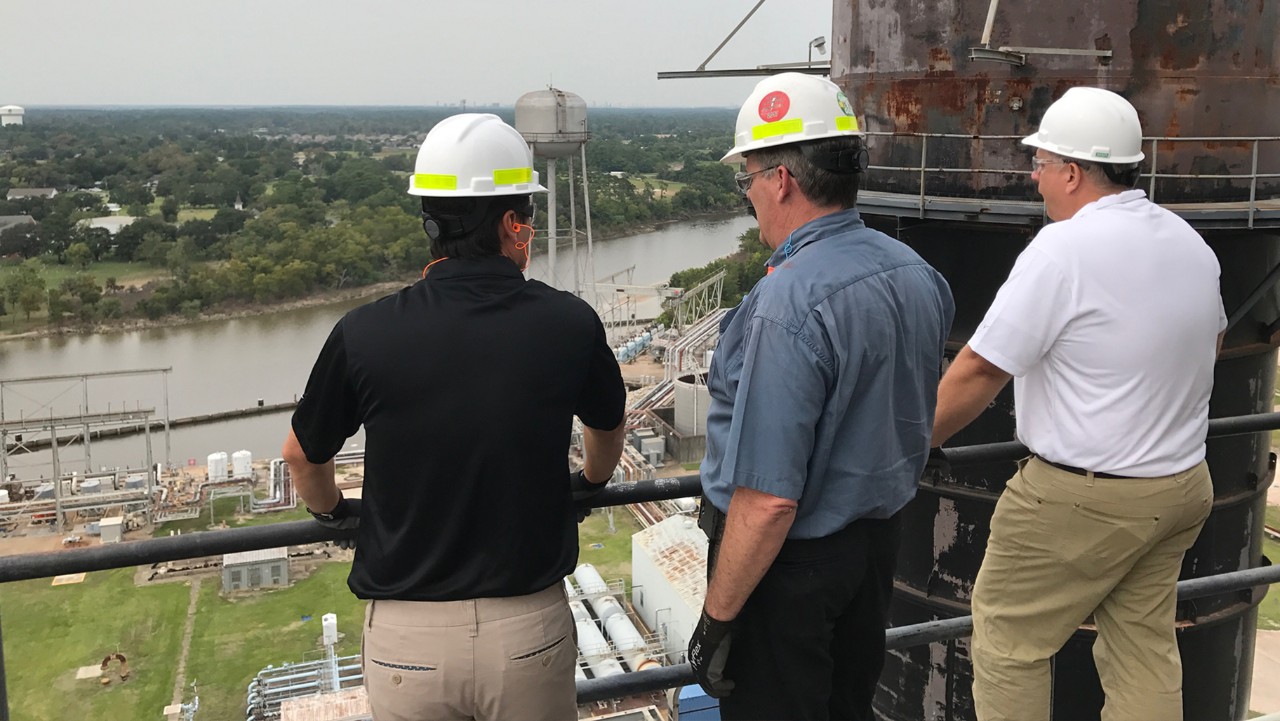 three men in hard hats stand on a platform, overlooking a river and industrial area with water towers.