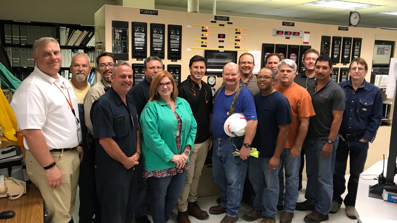 a group of people stand and smile together in a control room with panels and monitors in the background.