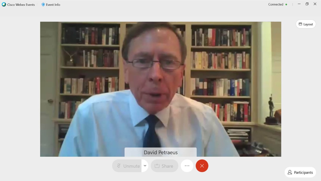 man in glasses and tie speaking during a video call, with bookshelves in the background.