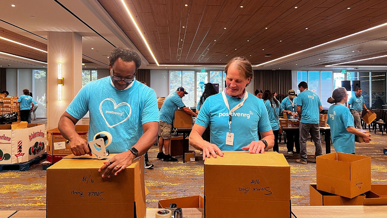 volunteers in blue shirts pack cardboard boxes in a large, well-lit room with tables and supplies.