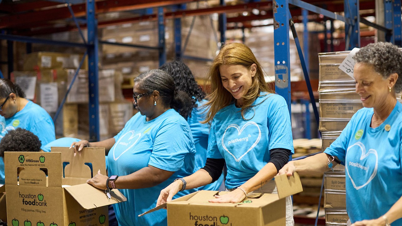 four women pack boxes at a food bank, all wearing blue shirts with a white heart design.