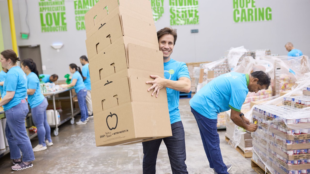 smiling man carries stacked boxes at a food bank, while others sort items and pack food in the background.