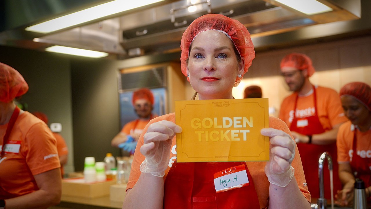 woman in red hairnet and gloves holds a Golden Ticket in a kitchen with other people in orange shirts.