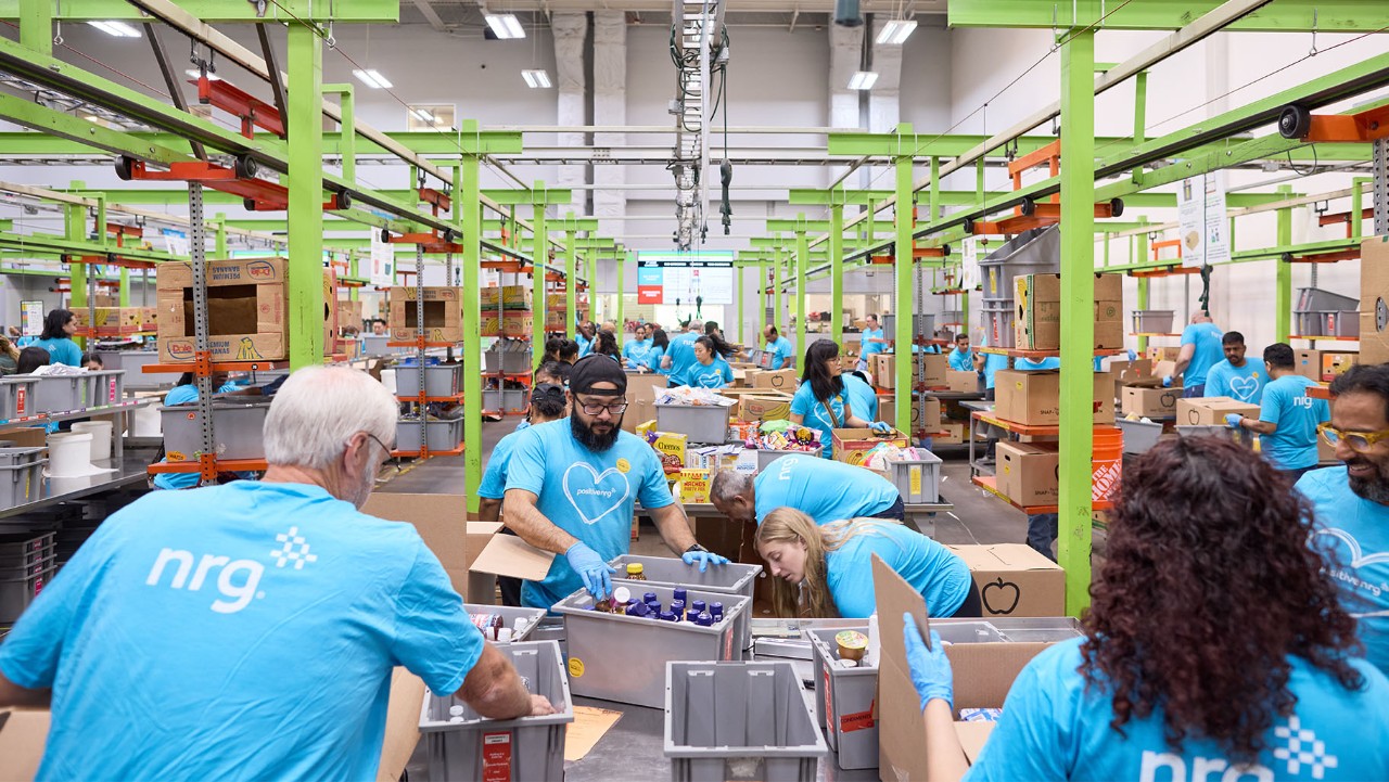 volunteers in blue shirts pack boxes of food in a brightly lit warehouse with green beams.