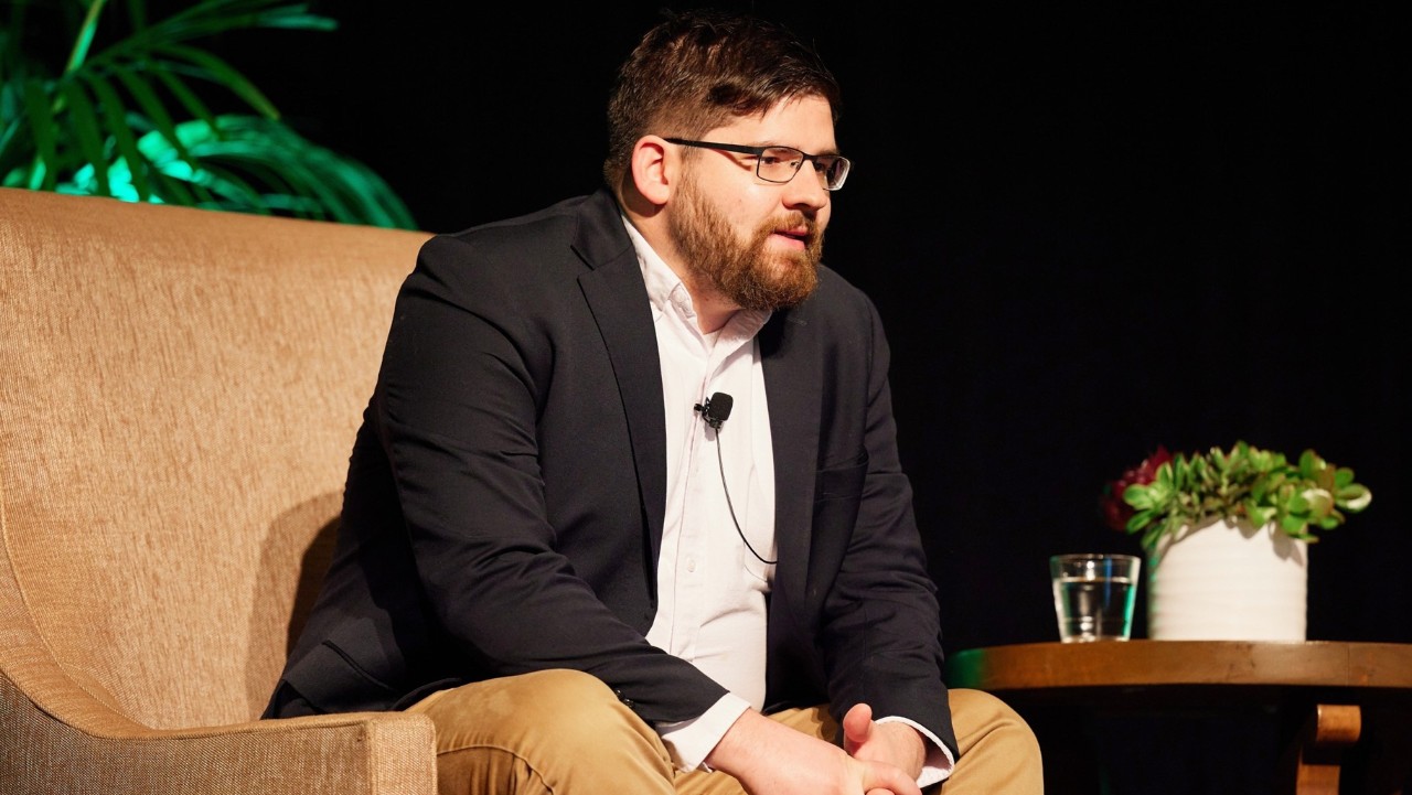 man in glasses sits on a brown couch, talking, with a plant and glass of water on a table nearby.