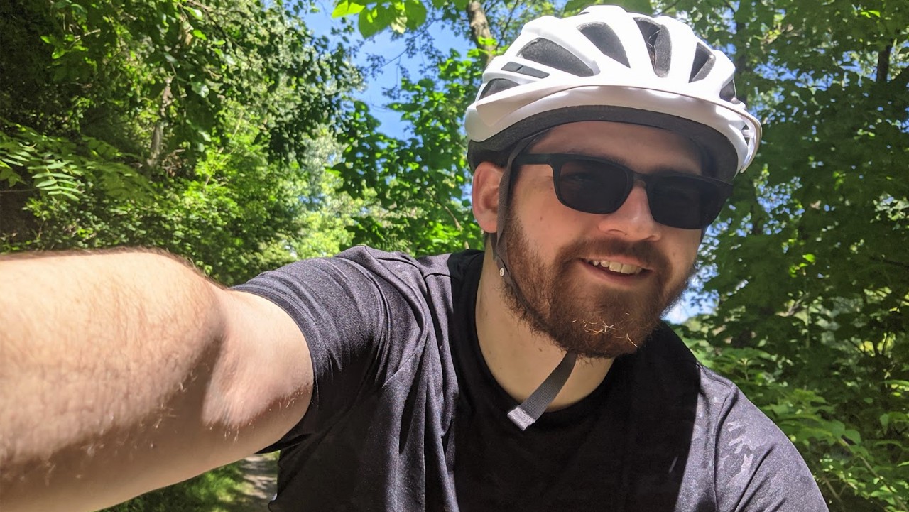bearded man in a bike helmet and sunglasses smiles on a sunny, tree-lined trail.