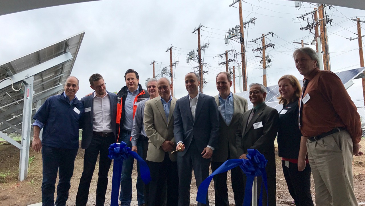 ten people stand together at a ribbon-cutting ceremony near solar panels and power lines outdoors.