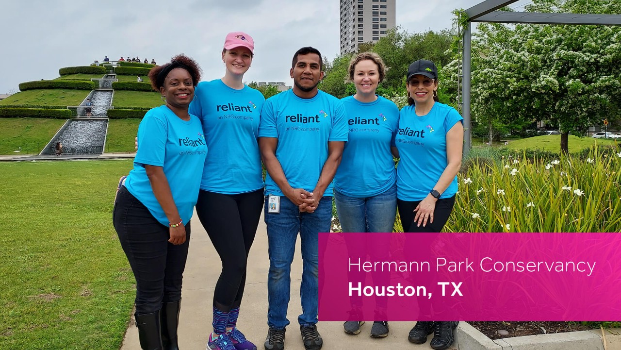 five people in blue shirts pose and smile at Hermann Park Conservancy in Houston, TX, with greenery behind them.