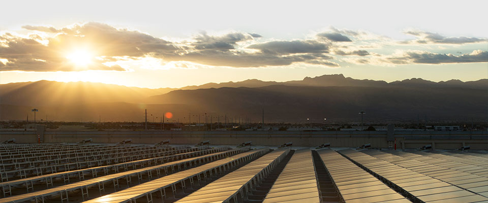 rows of solar panels reflect sunlight at sunset with mountains and clouds in the background.