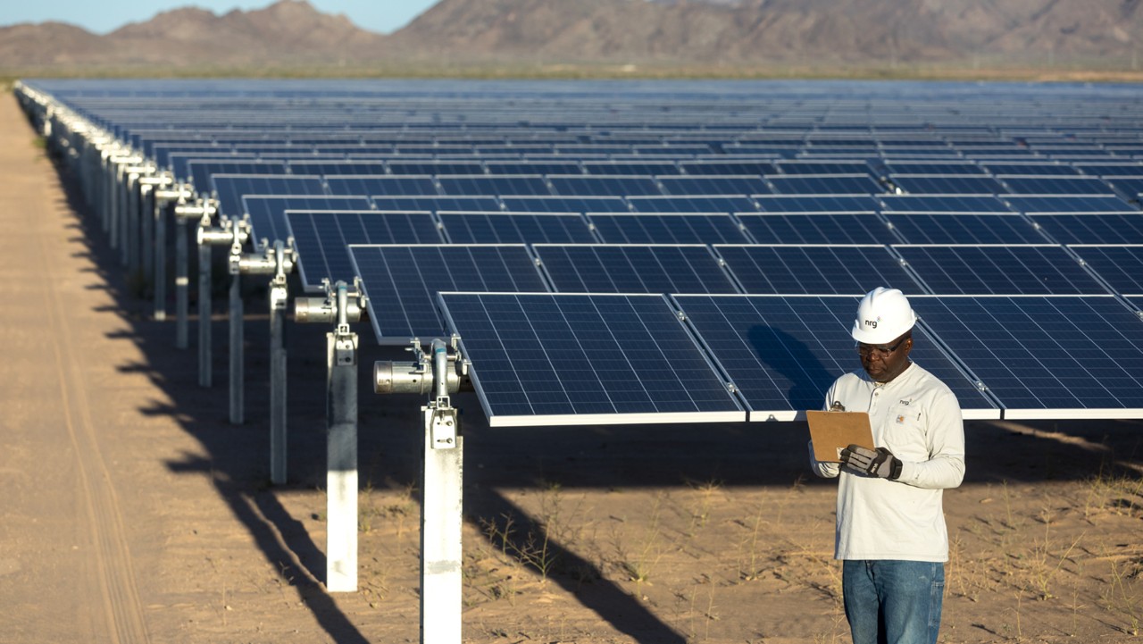man wearing a hard hat inspects solar panels in a large solar farm with mountains in the background.