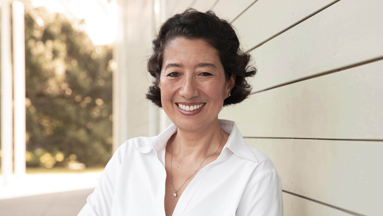 woman with short dark hair smiling, wearing a white shirt, standing by a light-colored wooden wall outdoors.