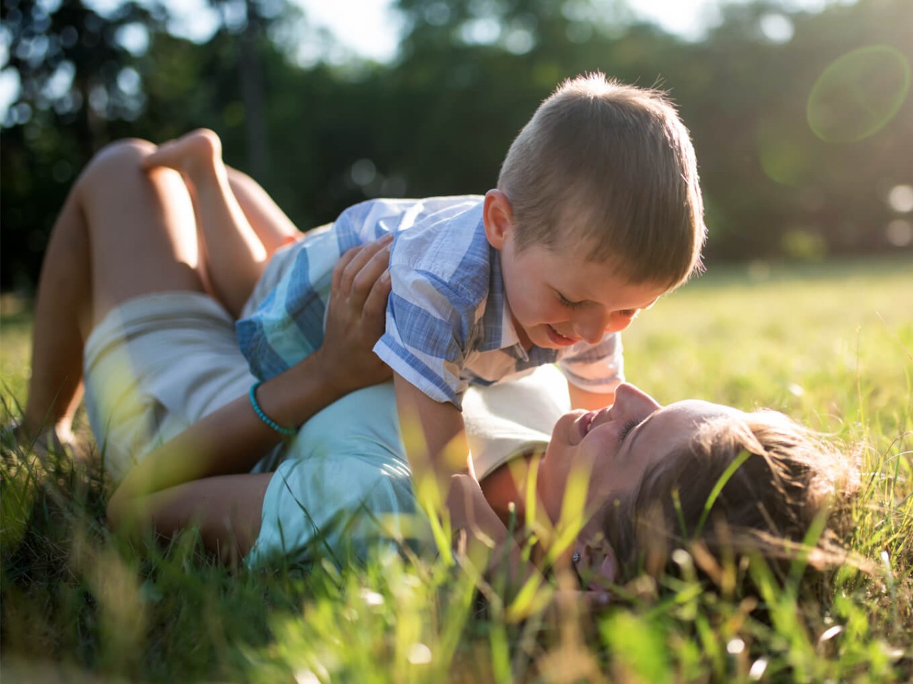 woman lying on grass, smiling, with a young boy on top of her, both laughing in the sunlight.
