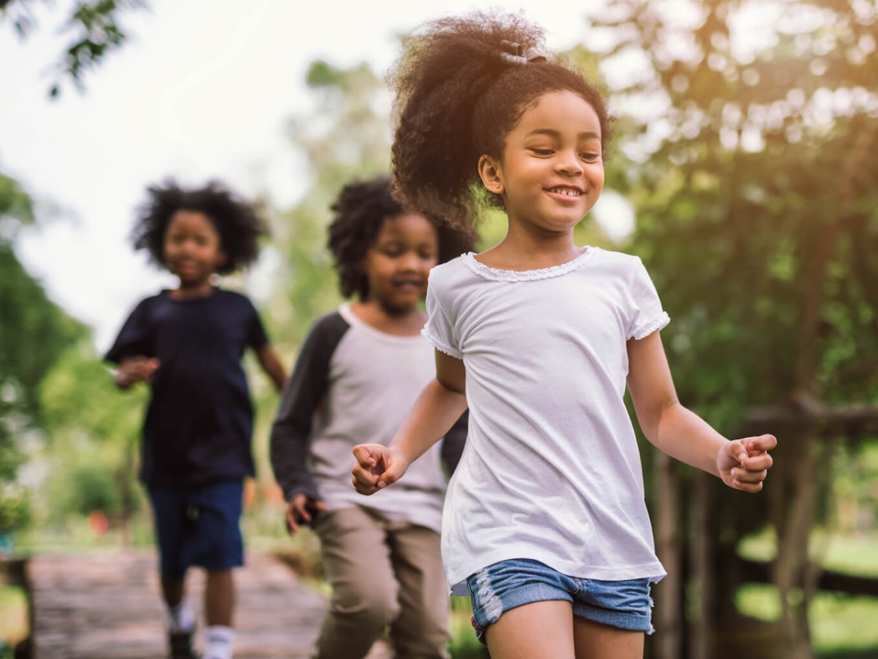 three children smiling and running outdoors on a sunny day, with trees and greenery in the background.