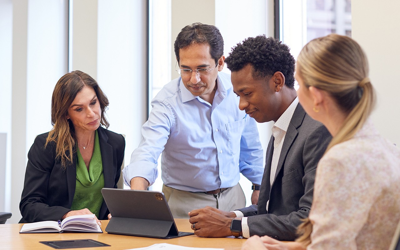 four people in business attire sit at a table, looking at a tablet together during a meeting.